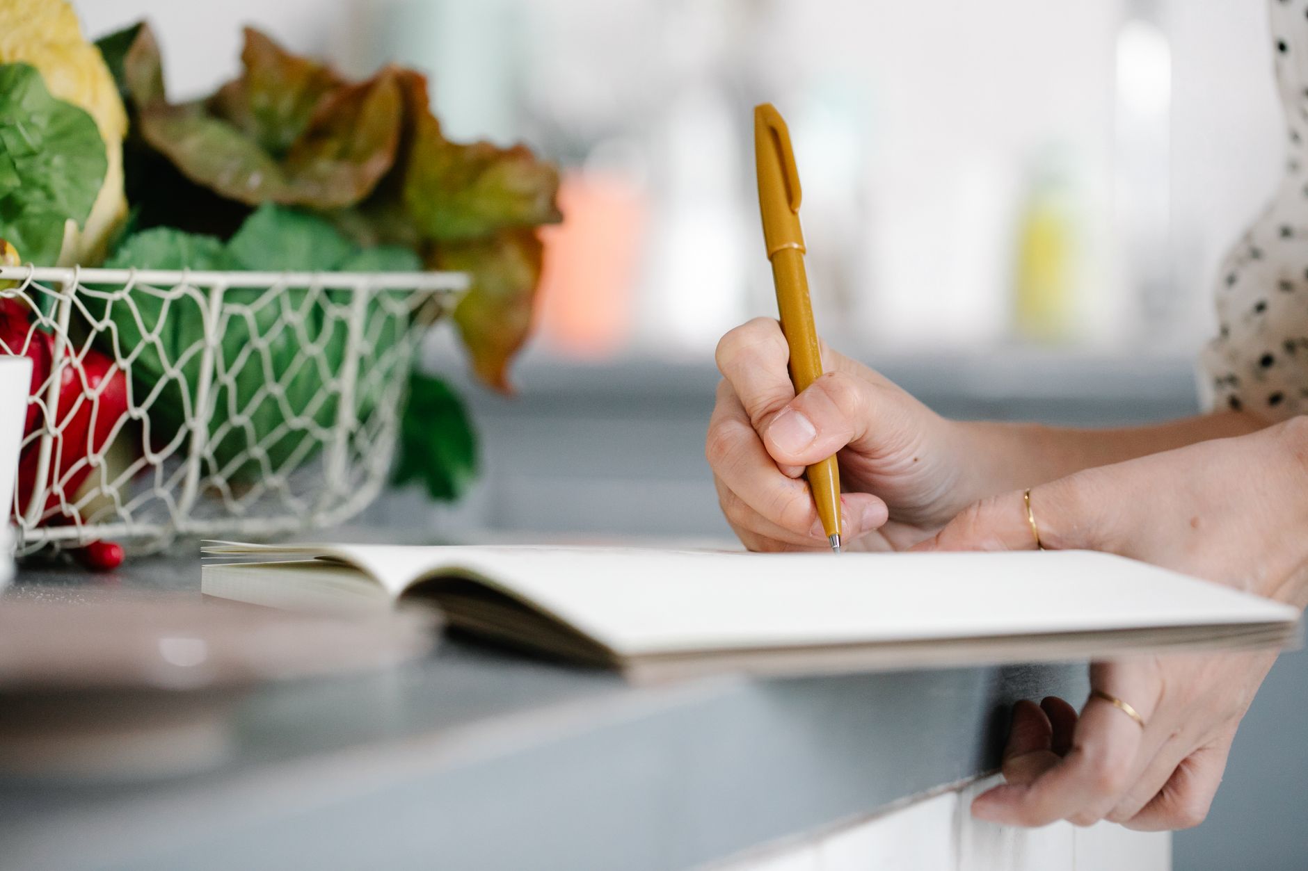 Frau schreibt mit einem Stift in ein Notizbuch, daneben ein Drahtkorb mit frischem Gemüse