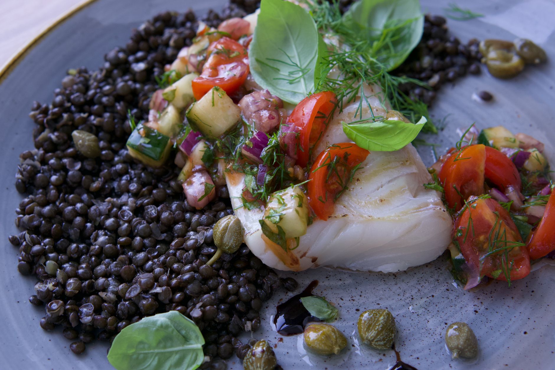 Weißes Fischfilet auf dunklen Linsen mit Tomaten, grünem Gemüse und frischem Basilikum auf einem grauen Teller