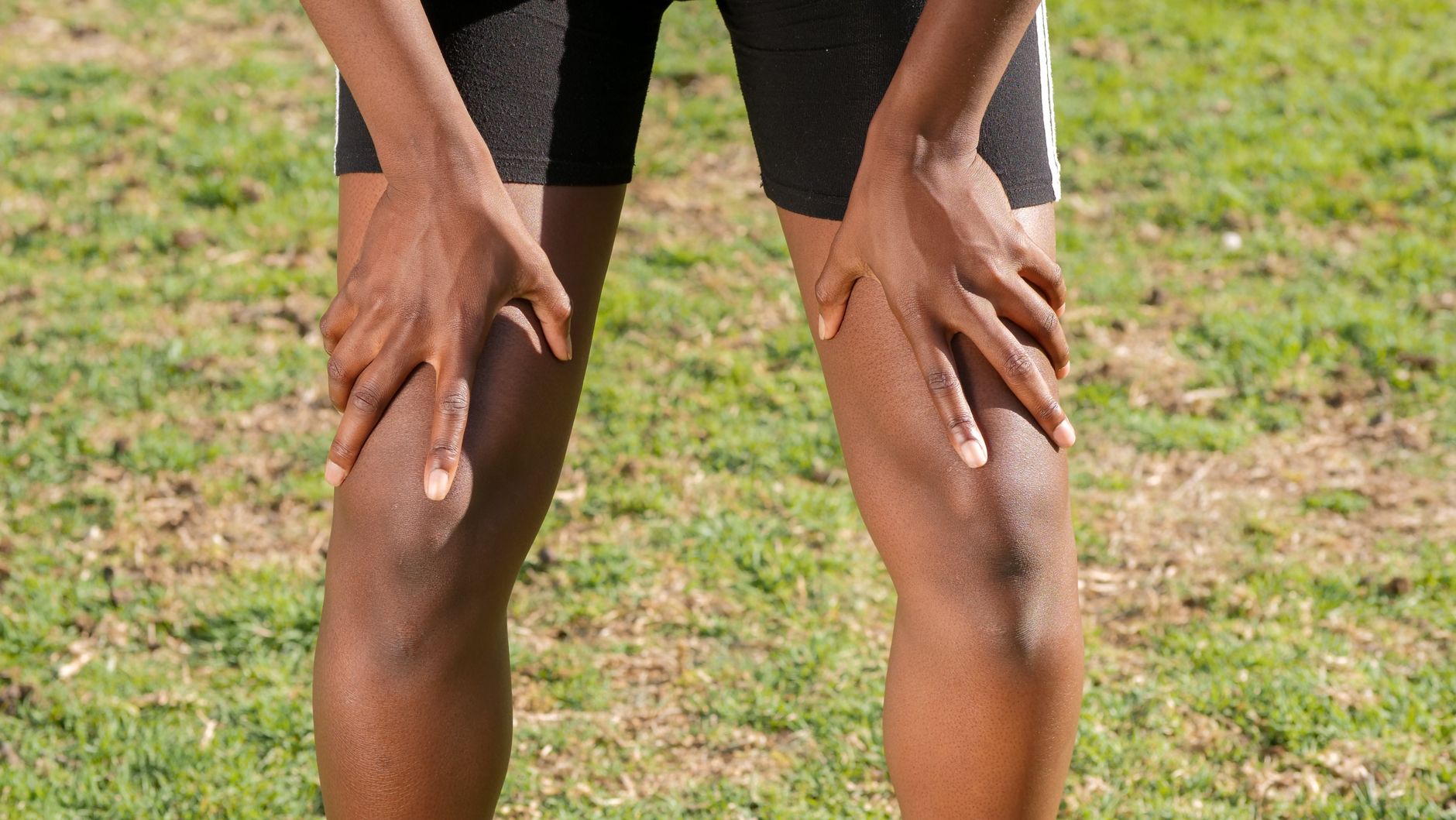 Person in Sportshorts stützt nach dem Training die Hände auf die Oberschenkel, Nahaufnahme auf einer Wiese
